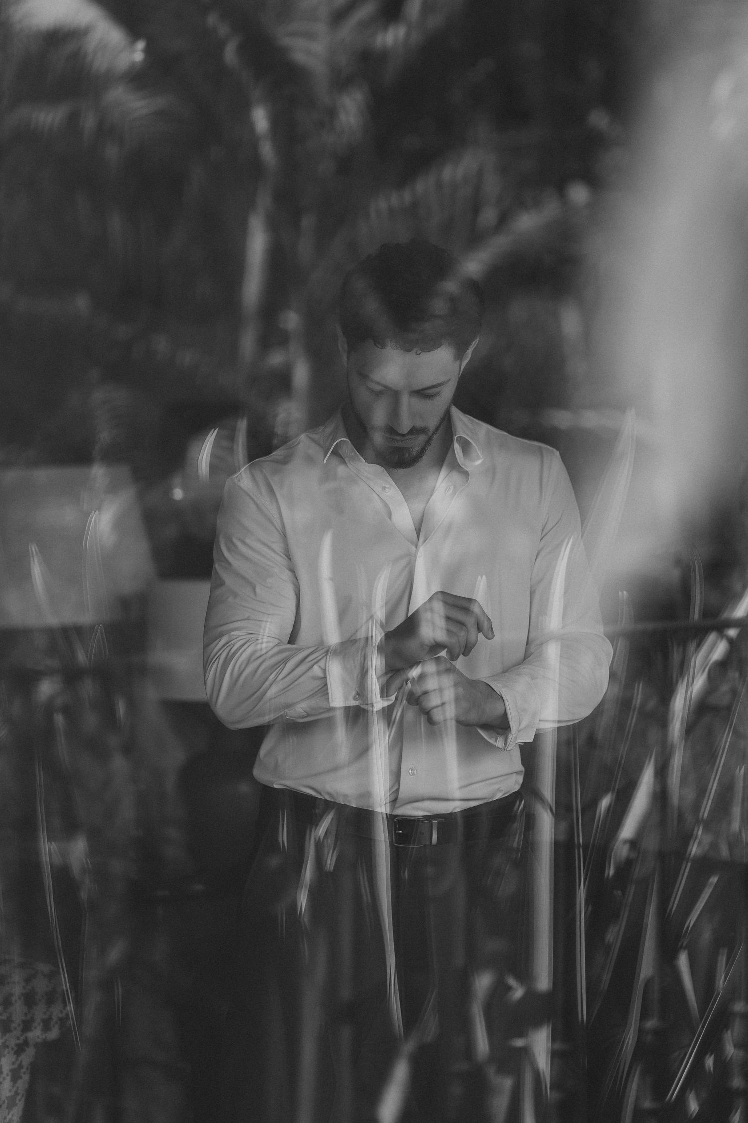 Artistic black and white portrait of person in white shirt looking down at wristwatch amidst foliage.