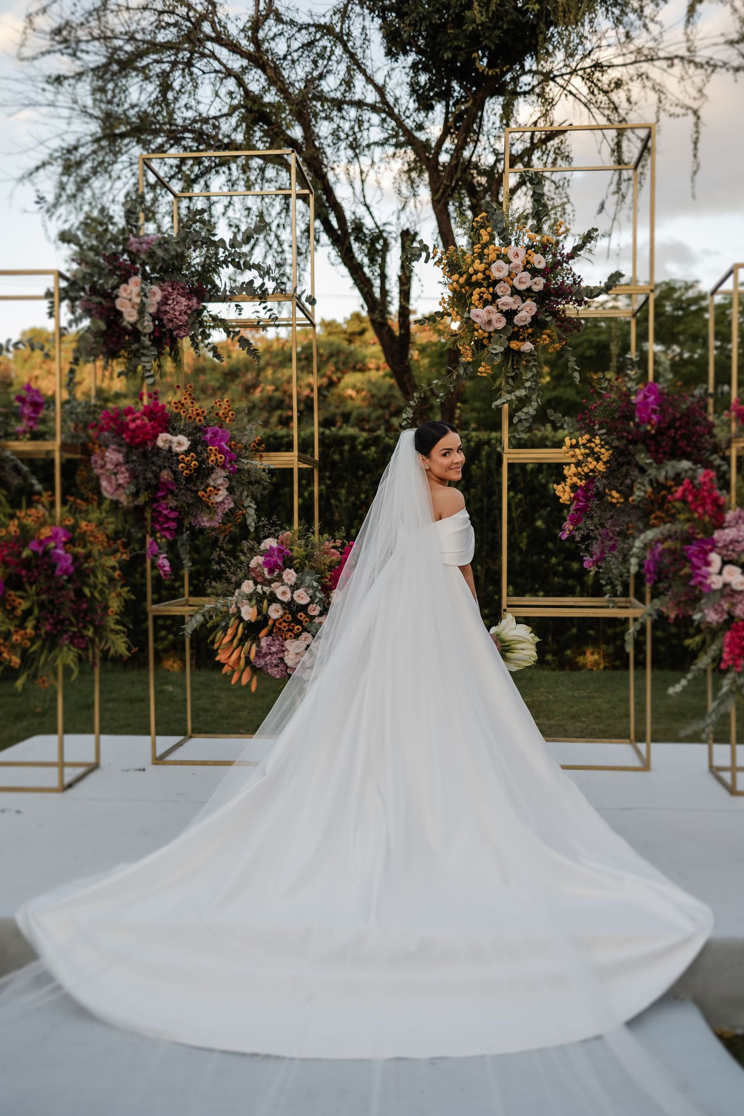 Beautiful bridal scene featuring flowing white wedding gown and floral arch decorated with purple and pink blooms outdoors.