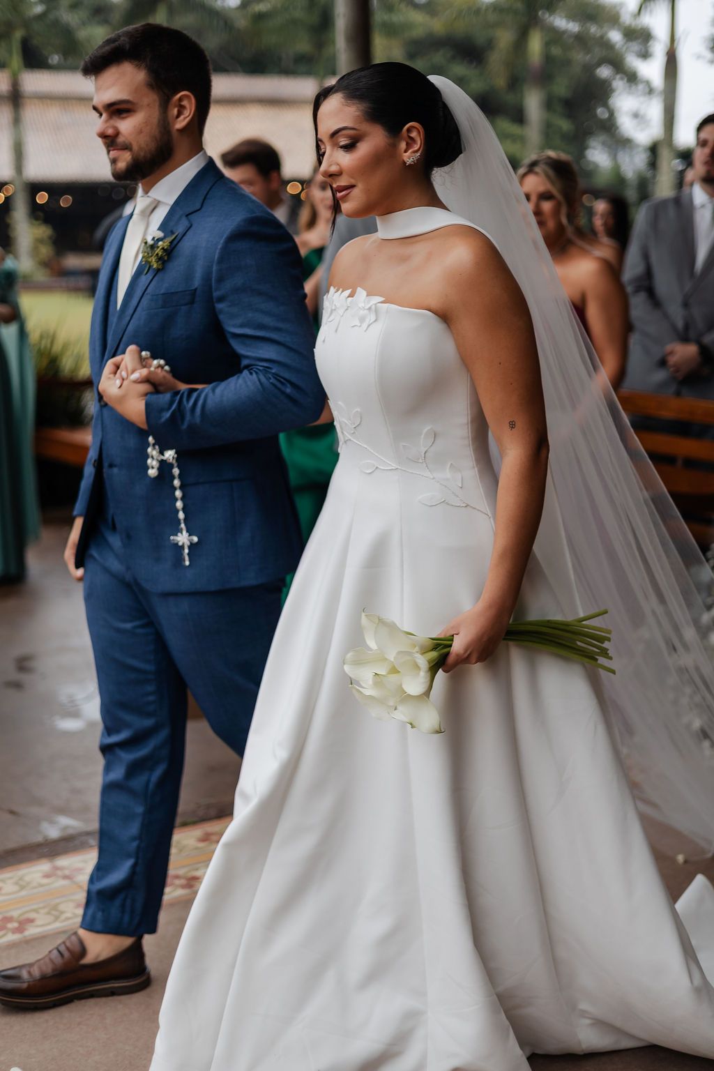 A couple at their wedding ceremony, with the bride in a white strapless gown holding calla lilies and groom in navy suit.