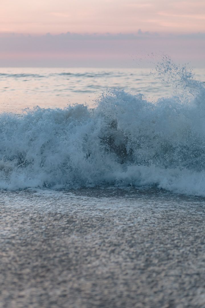 A powerful ocean wave crashes and sprays against the shoreline during a pink sunset.