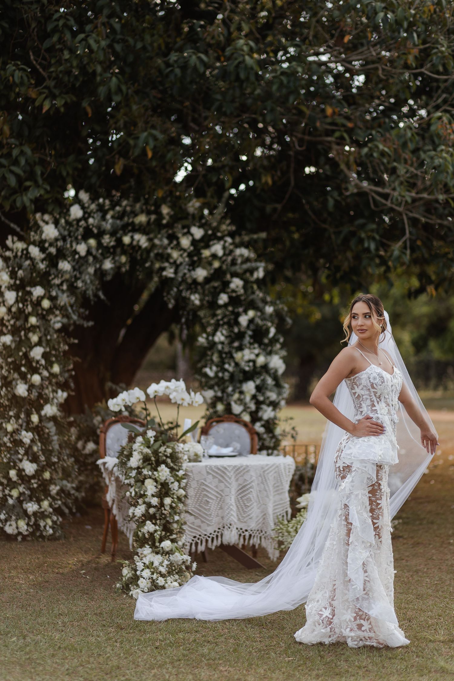 A bride in a flowing lace wedding dress poses gracefully under a blooming magnolia tree in an outdoor garden setting.