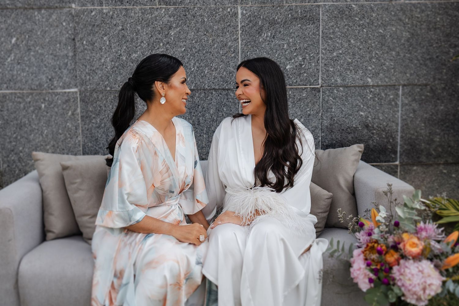 Two women in white robes share joyful moments while sitting on a grey sofa with colorful flowers nearby.