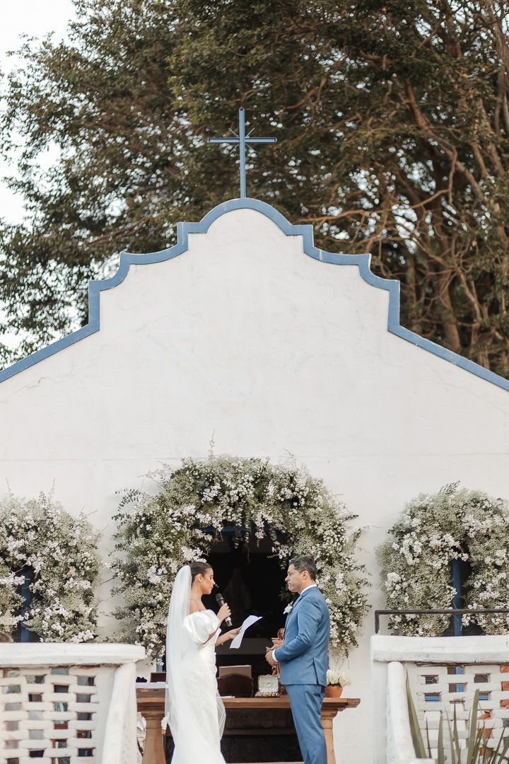 A couple stands at a white chapel altar under an arch adorned with flowers during their wedding ceremony.