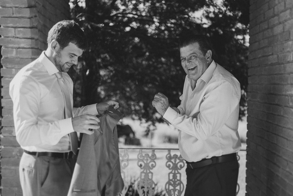 Two men in light-colored shirts laugh together while standing on a porch near a brick wall.