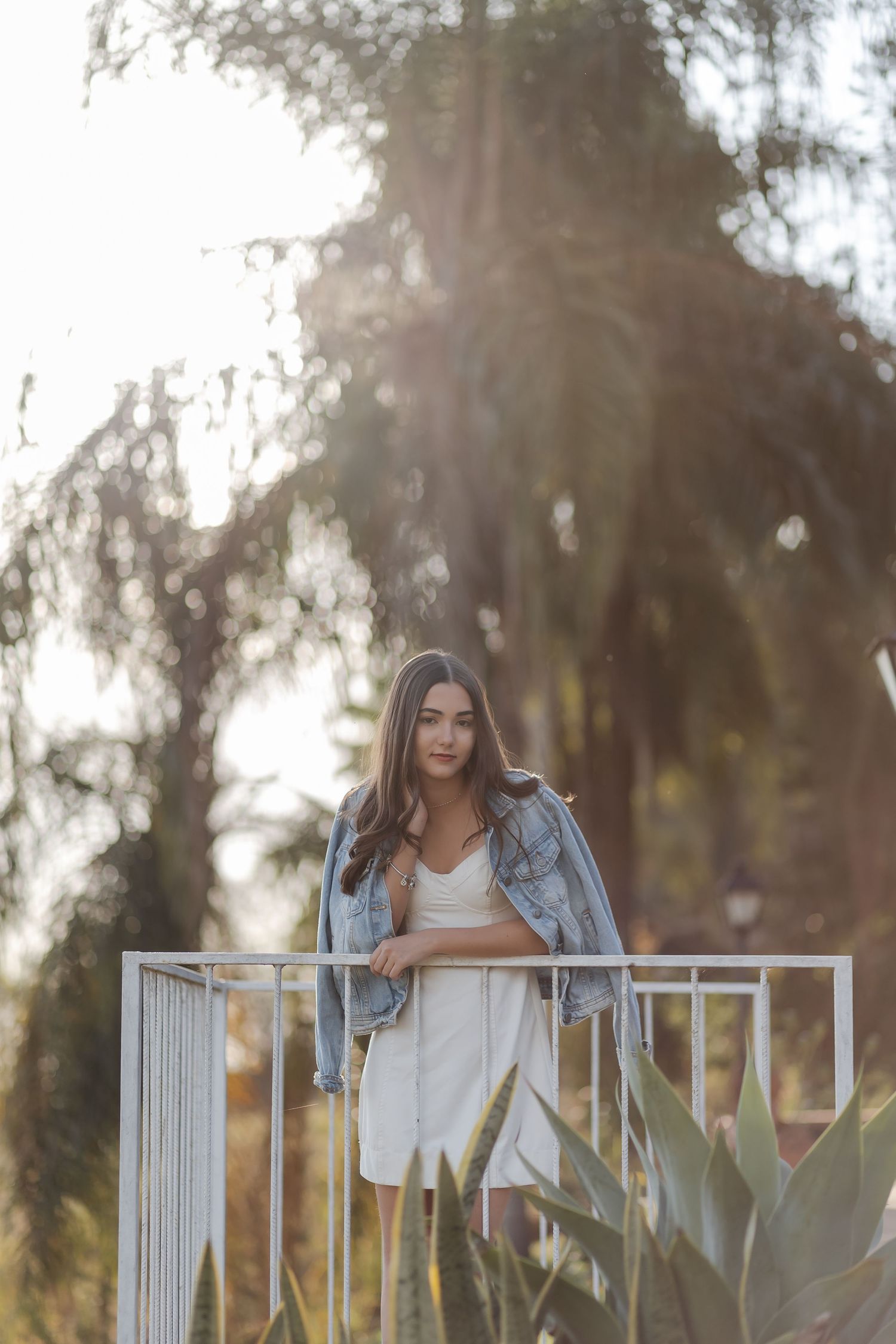 A woman in a white top and denim jacket leans on a white railing in a sunny outdoor setting with trees.
