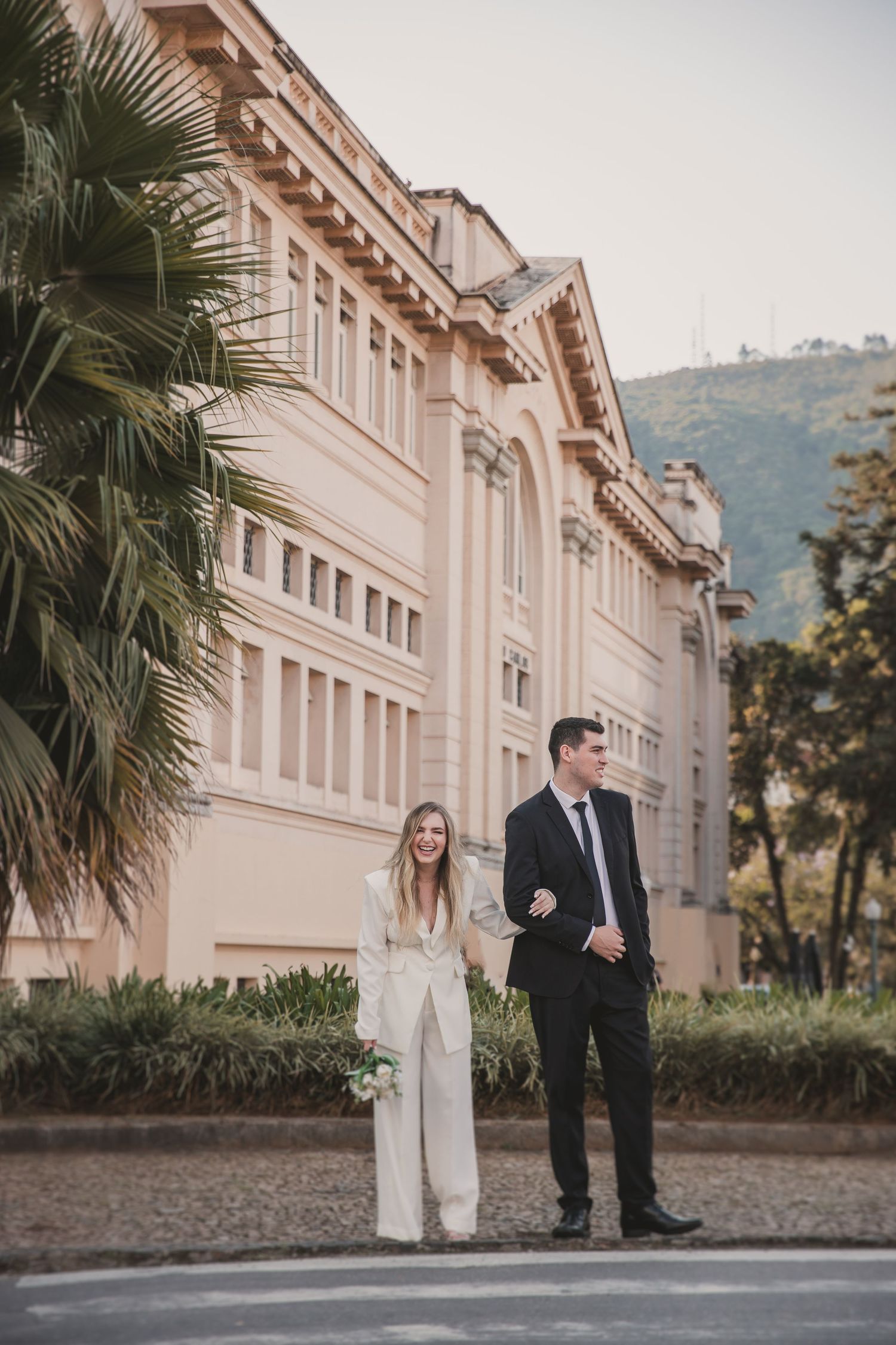 A couple stands elegantly in front of a grand beige historic building with palm trees and mountain views in the background.