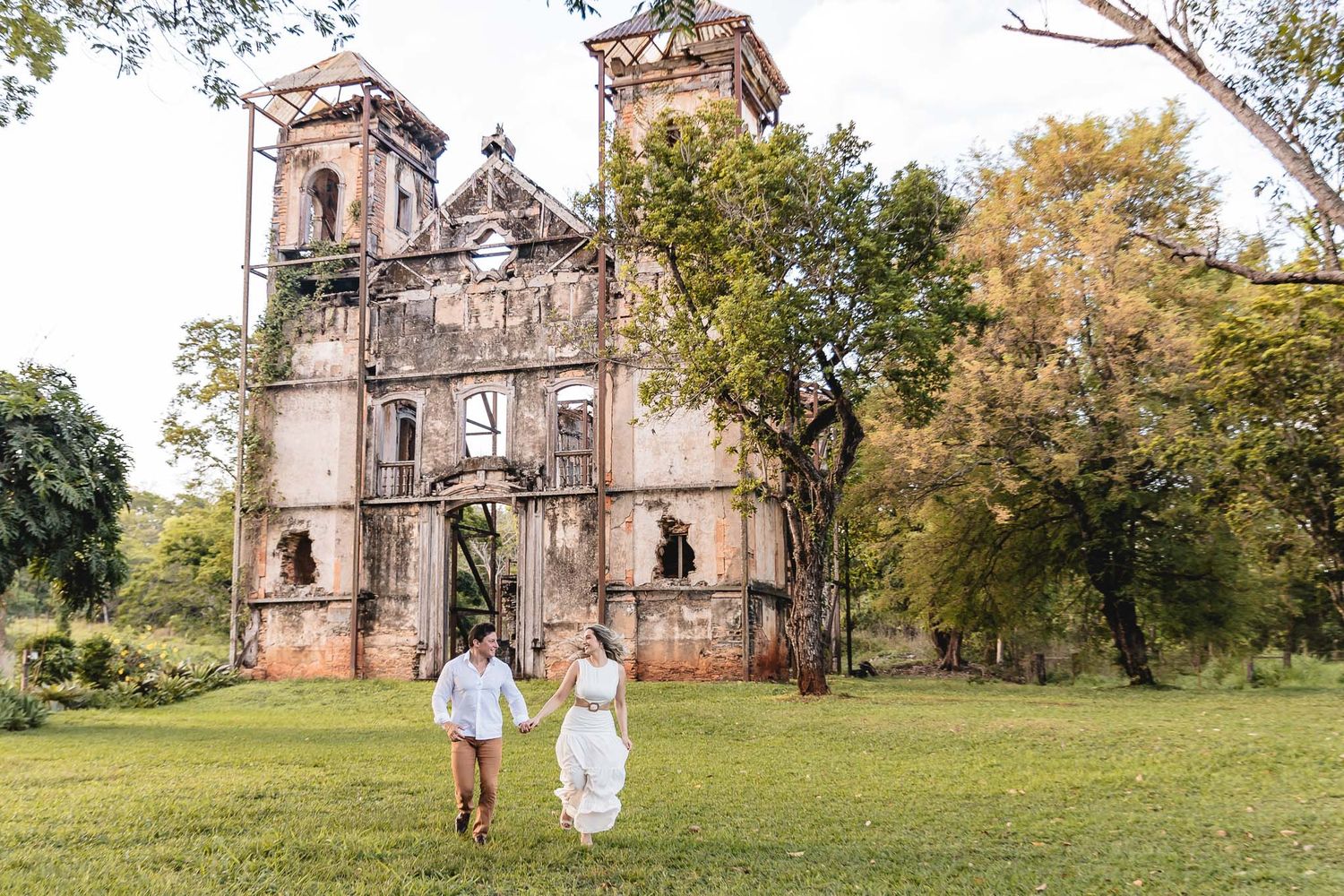 Two figures in light clothing run hand in hand across a grassy lawn in front of a weathered historic church with twin towers.