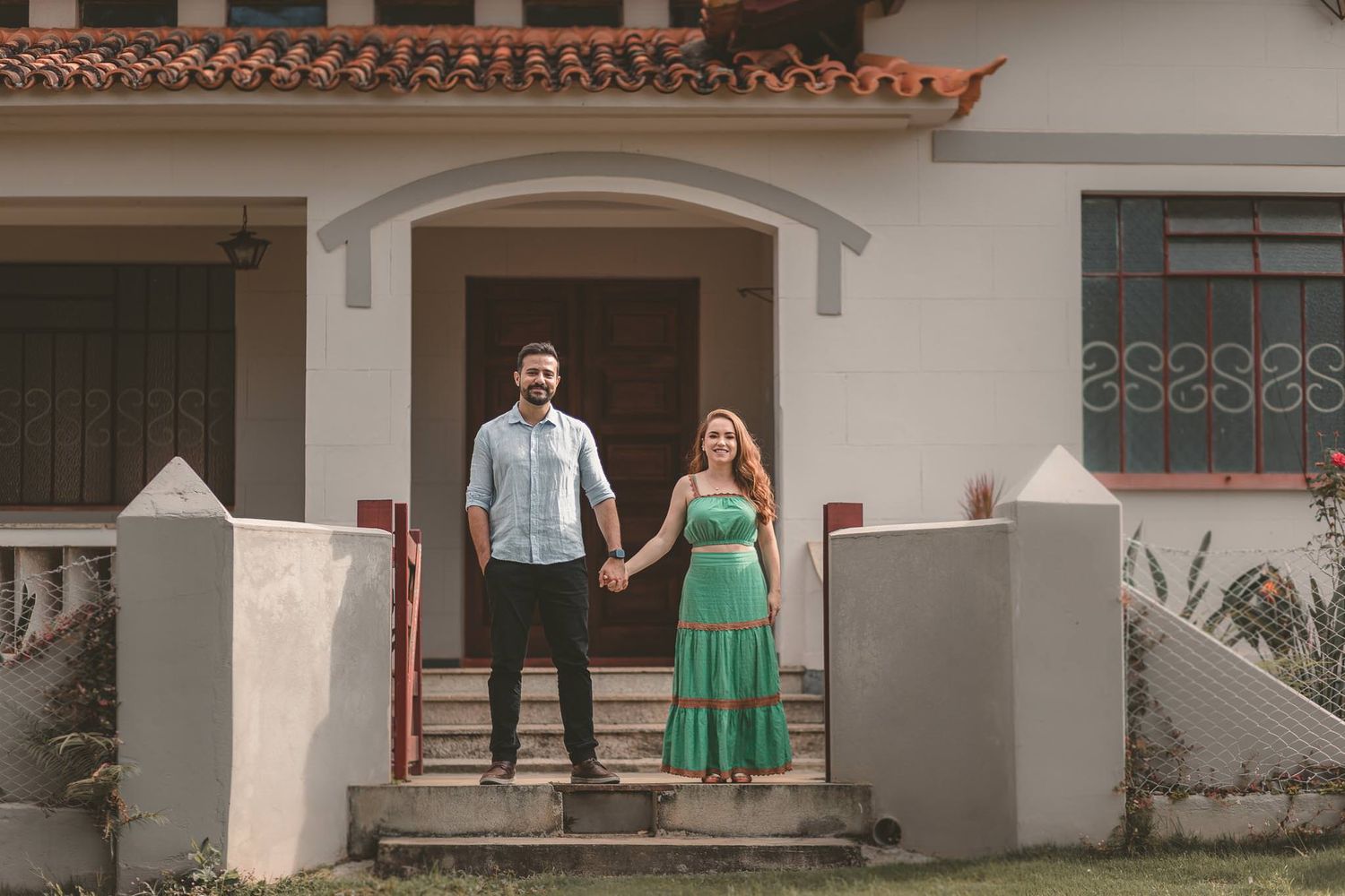 A couple stands together holding hands on the front steps of a Mediterranean-style home with terracotta roof tiles.