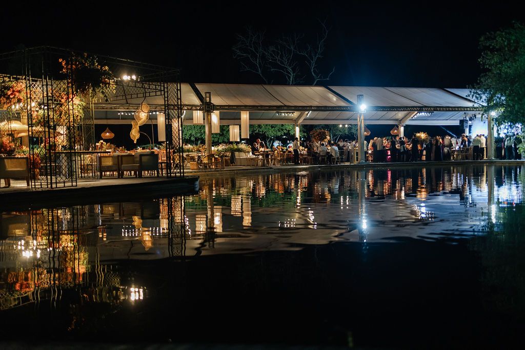 An illuminated waterfront restaurant with reflective pool at night showing warm lights and diners under covered pavilions.