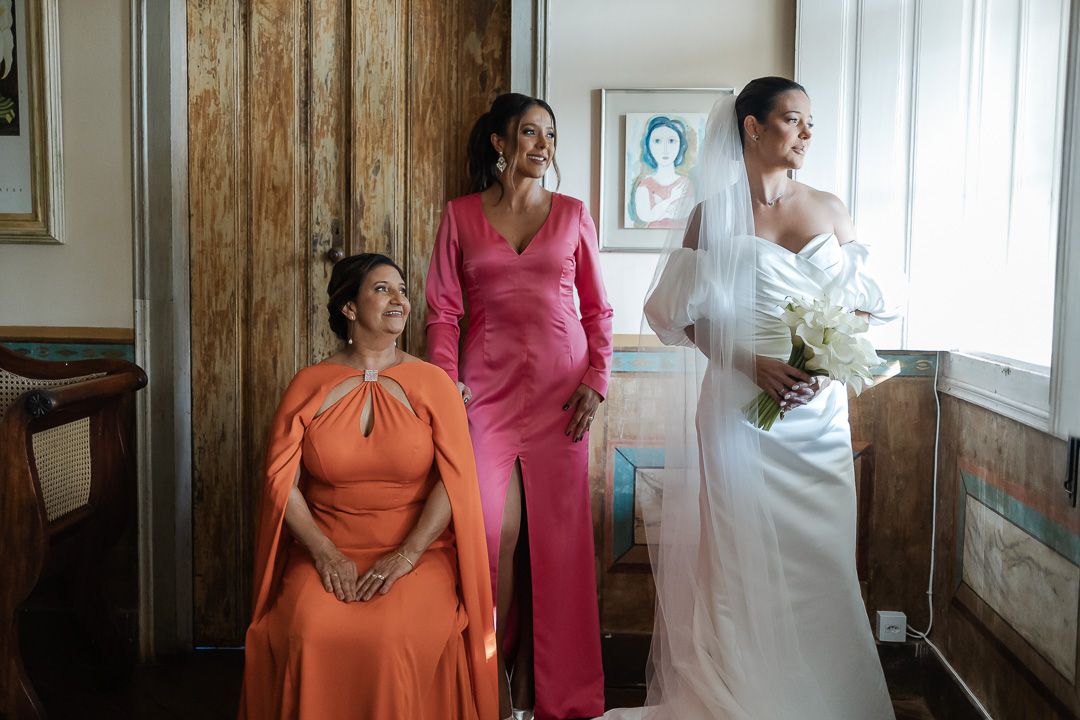 Three women in formal dresses including orange, pink, and white gowns pose together in an elegant indoor setting.
