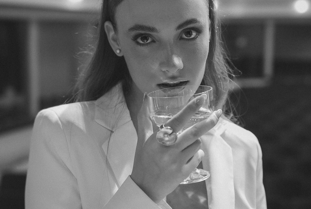 Black and white portrait of someone in white blazer holding cocktail glass with dramatic lighting and intense gaze.
