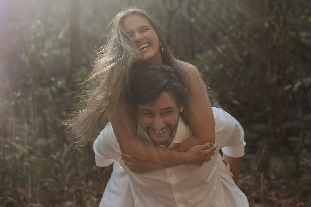 A joyful couple in white clothing share a playful piggyback ride moment in a sunlit forest setting.