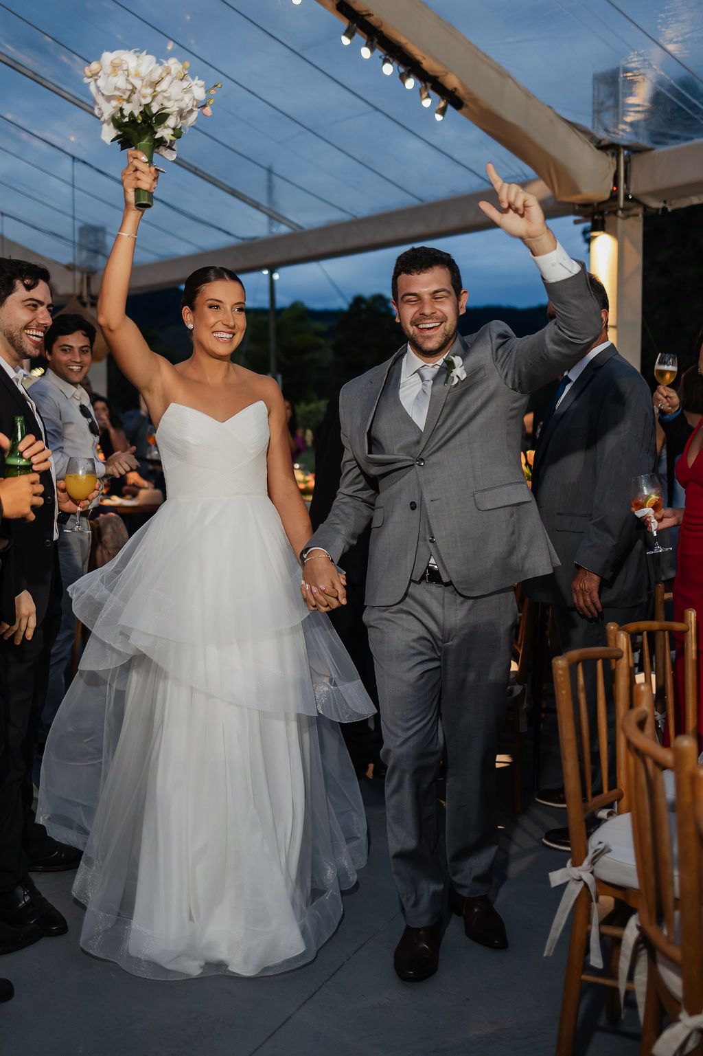 Newlyweds celebrate joyfully as they exit their wedding ceremony under string lights at dusk.