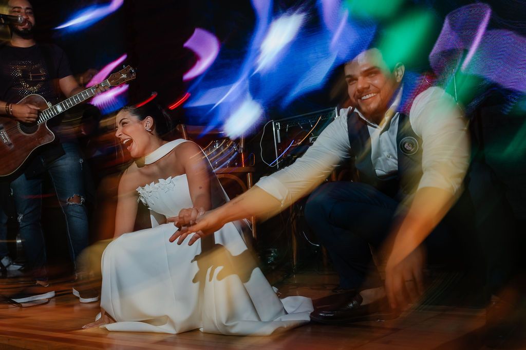 Wedding couple dances energetically on the dance floor with colorful light trails creating motion blur effect in background.