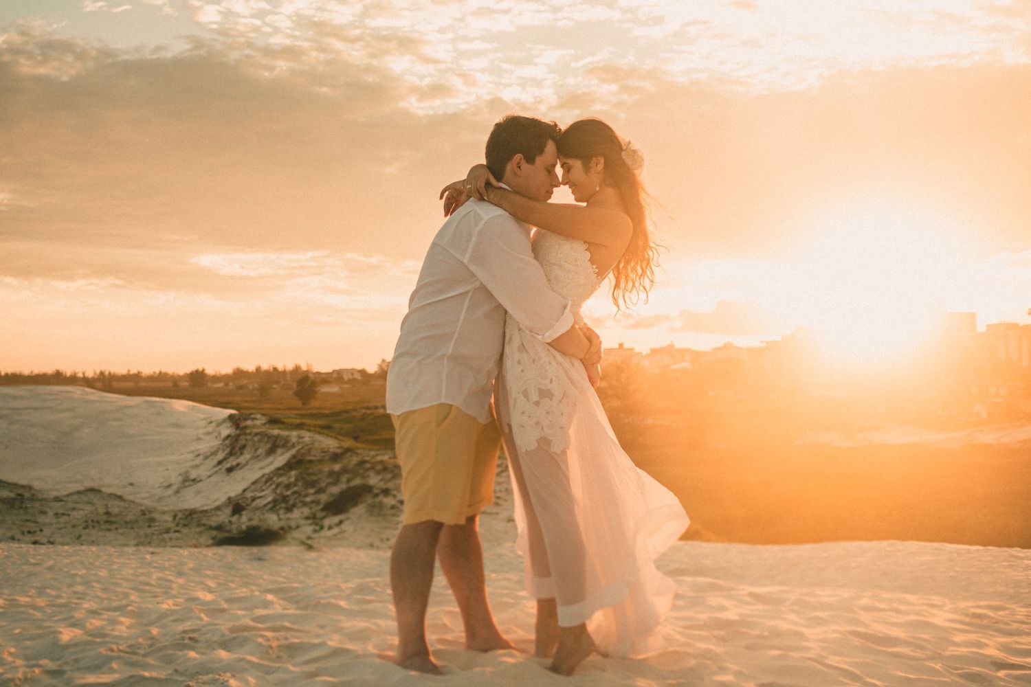 A romantic couple embraces on sandy dunes during a golden sunset, silhouetted against the warm glowing sky.