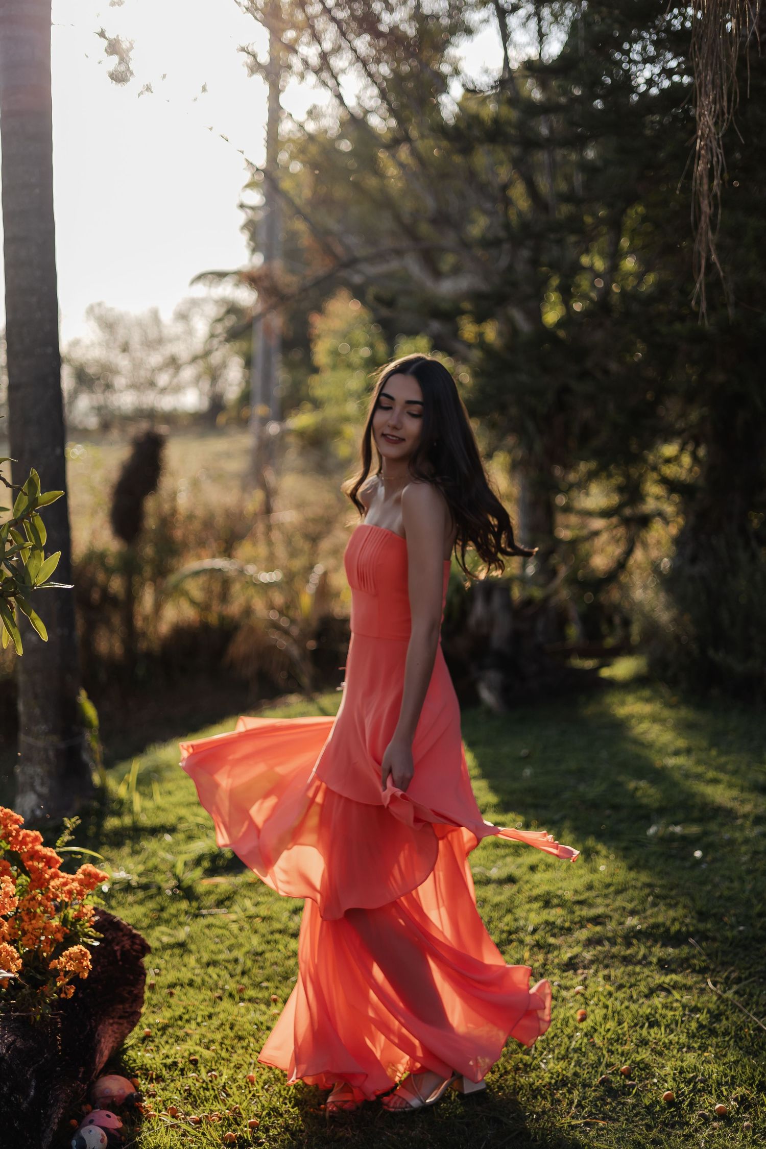 Person in a flowing coral dress twirls gracefully among trees in golden sunlight during an outdoor photoshoot.
