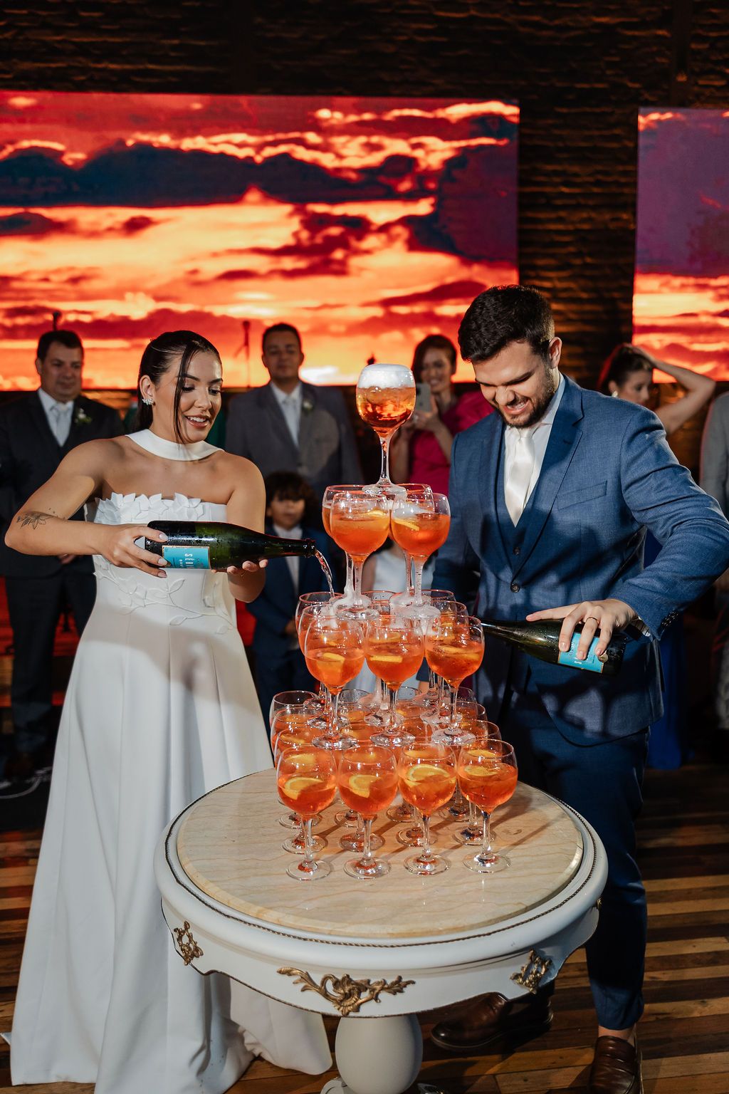 A couple pouring champagne into a tower of cocktail glasses against a dramatic sunset backdrop at their celebration.