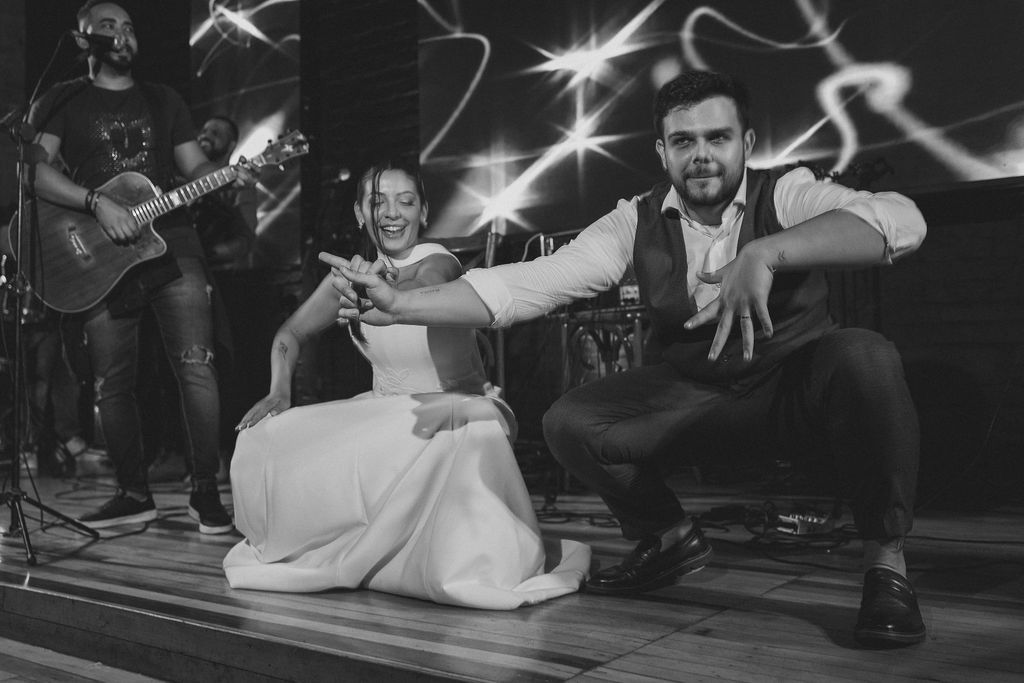 Black and white photo of newlyweds performing a playful dance move on a wooden floor with live band in background.
