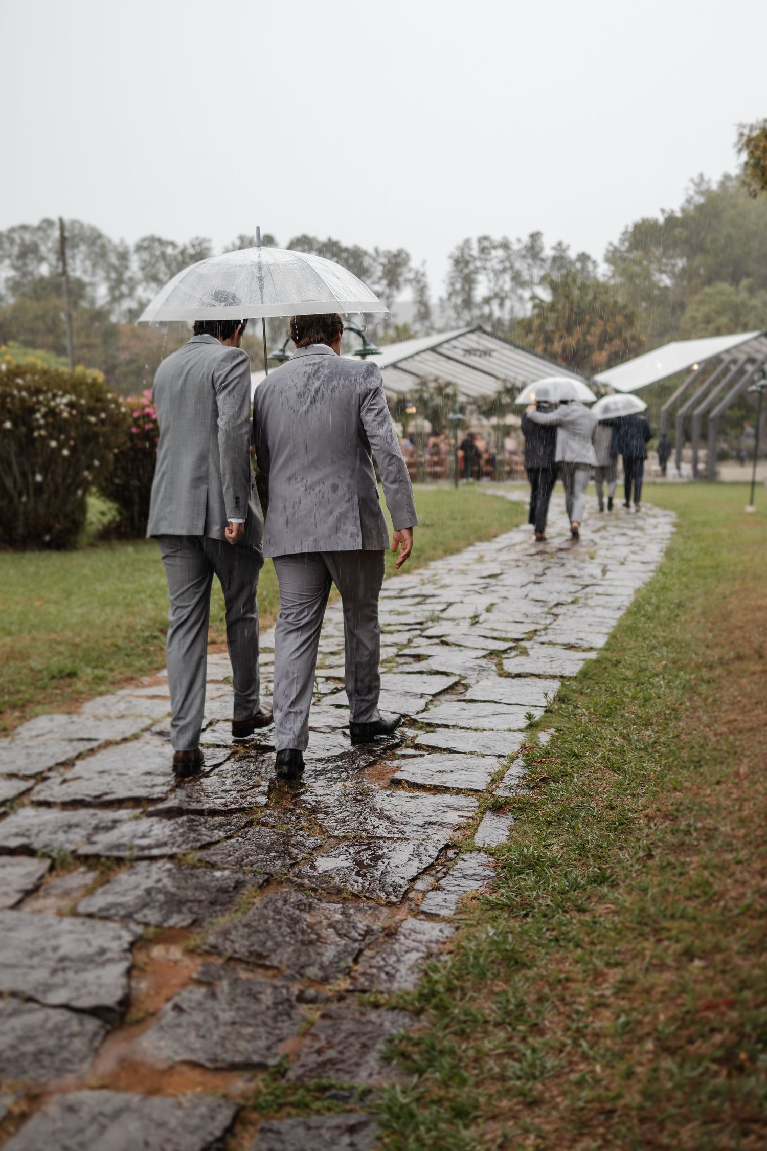 Wedding party members in matching gray suits walk along a wet stone path carrying umbrellas on a rainy day.