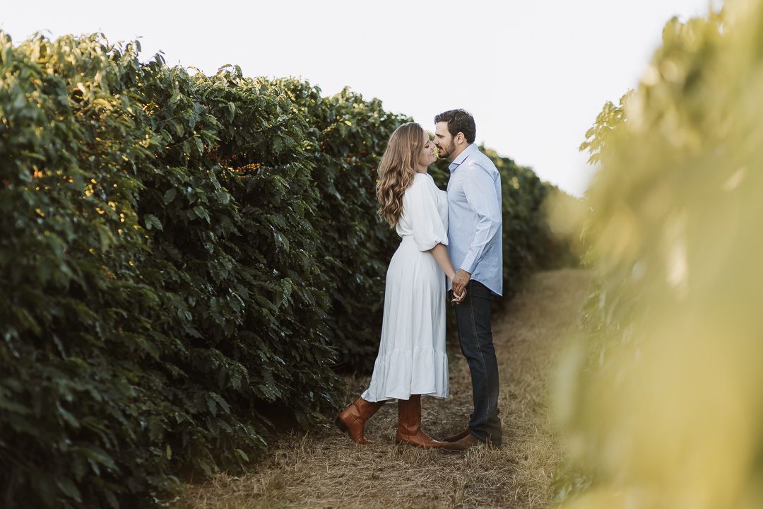 A couple stands together on a dirt path next to a tall hedge wall during golden hour.