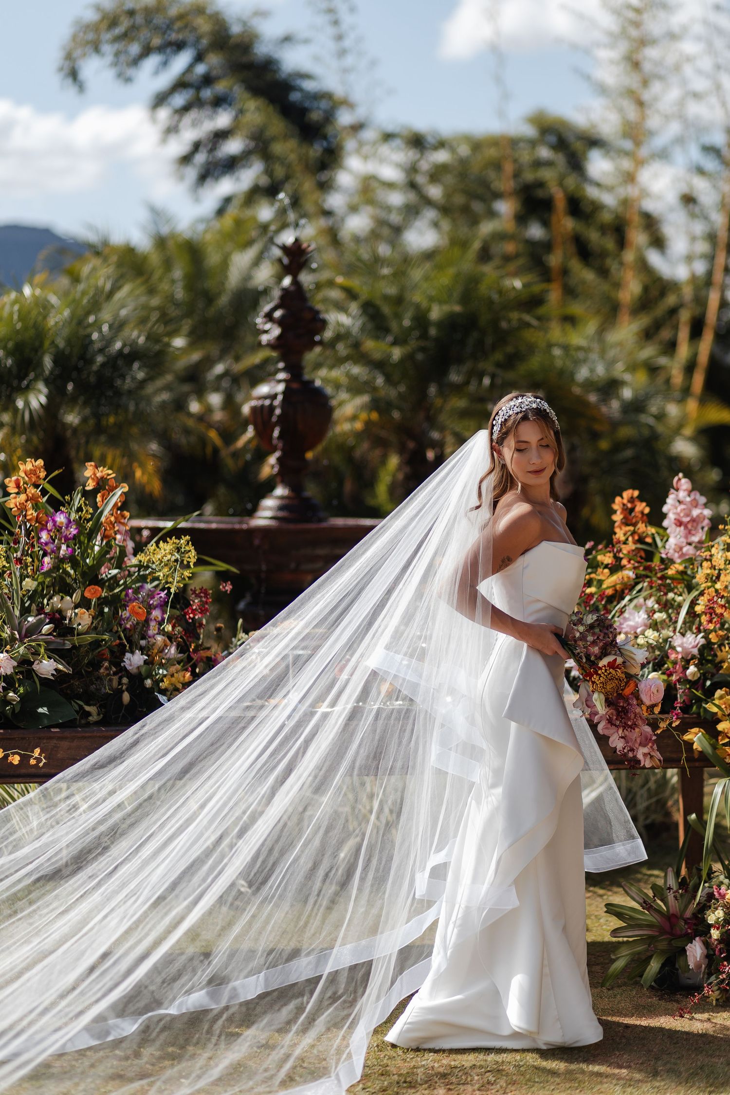 A bride in a elegant white dress with a flowing cathedral veil poses in a garden setting with colorful flowers at sunset.
