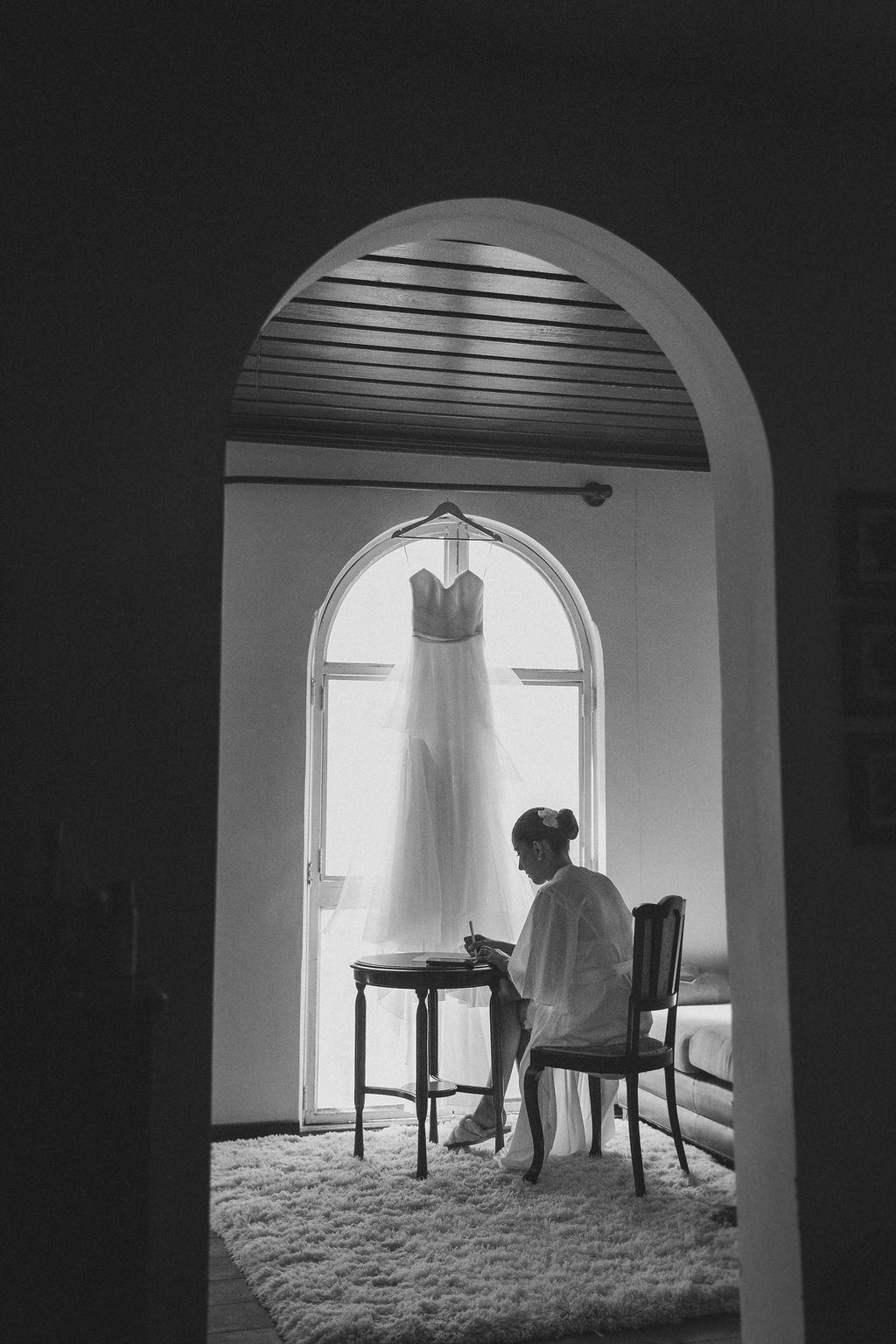 Cerimônia de casamento ao ar livre na FA black and white photo through an archway shows a white dress hanging while someone sits at a table.