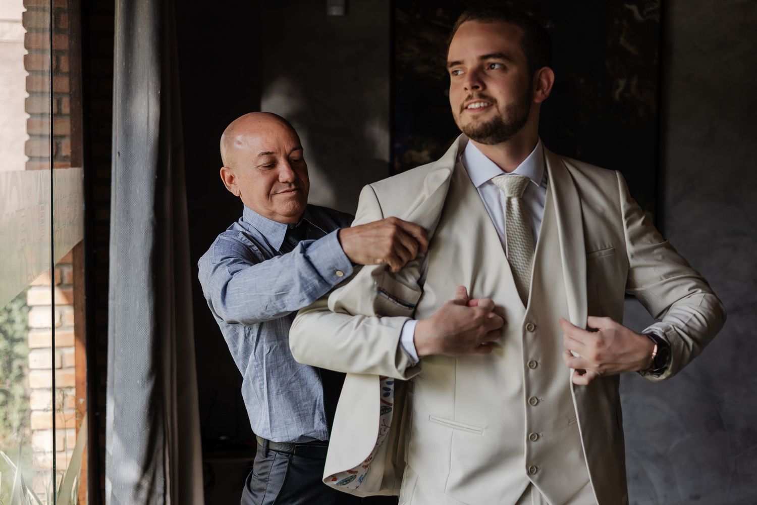 Man helping adjust the lapel of a light beige suit jacket in a dimly lit room with brick walls.