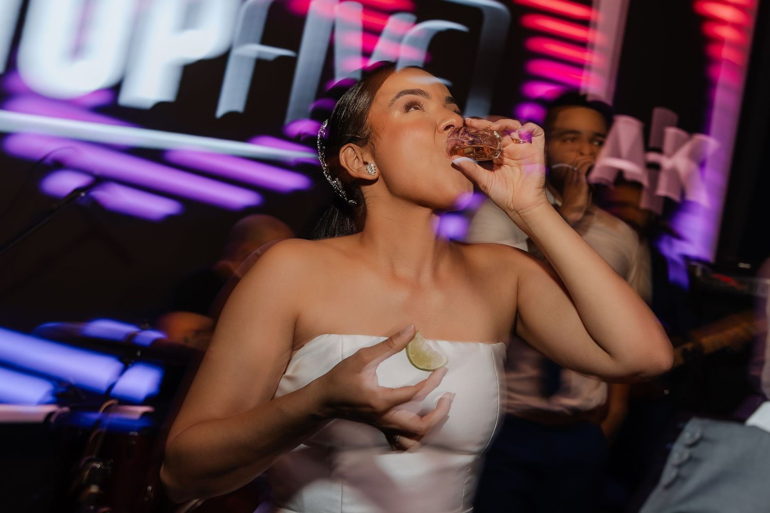 A person in a white strapless dress drinks from a small glass at a nightclub with colorful neon lights in the background.