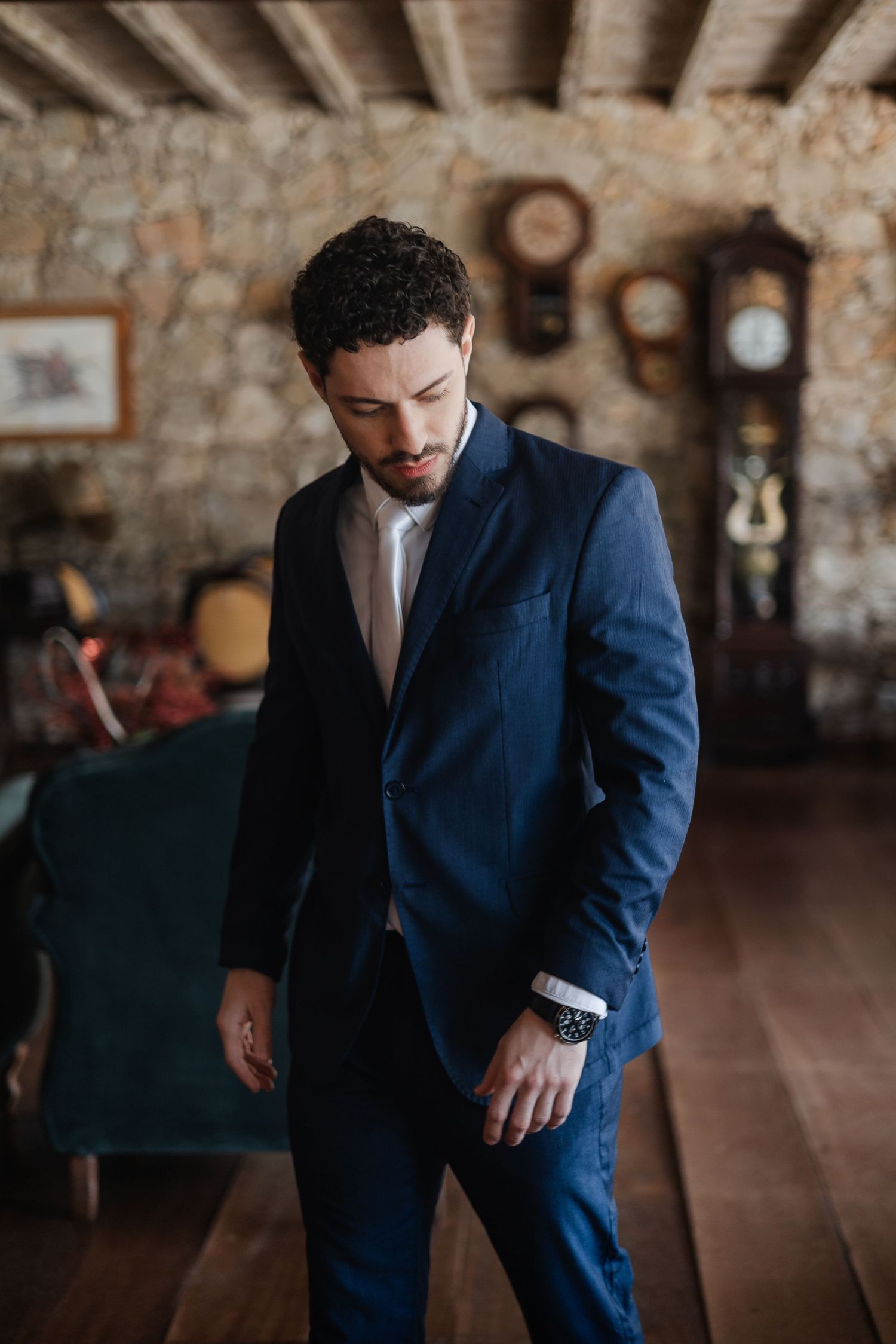A person in a navy blue suit adjusts their sleeve in a rustic room with stone walls and vintage decor.