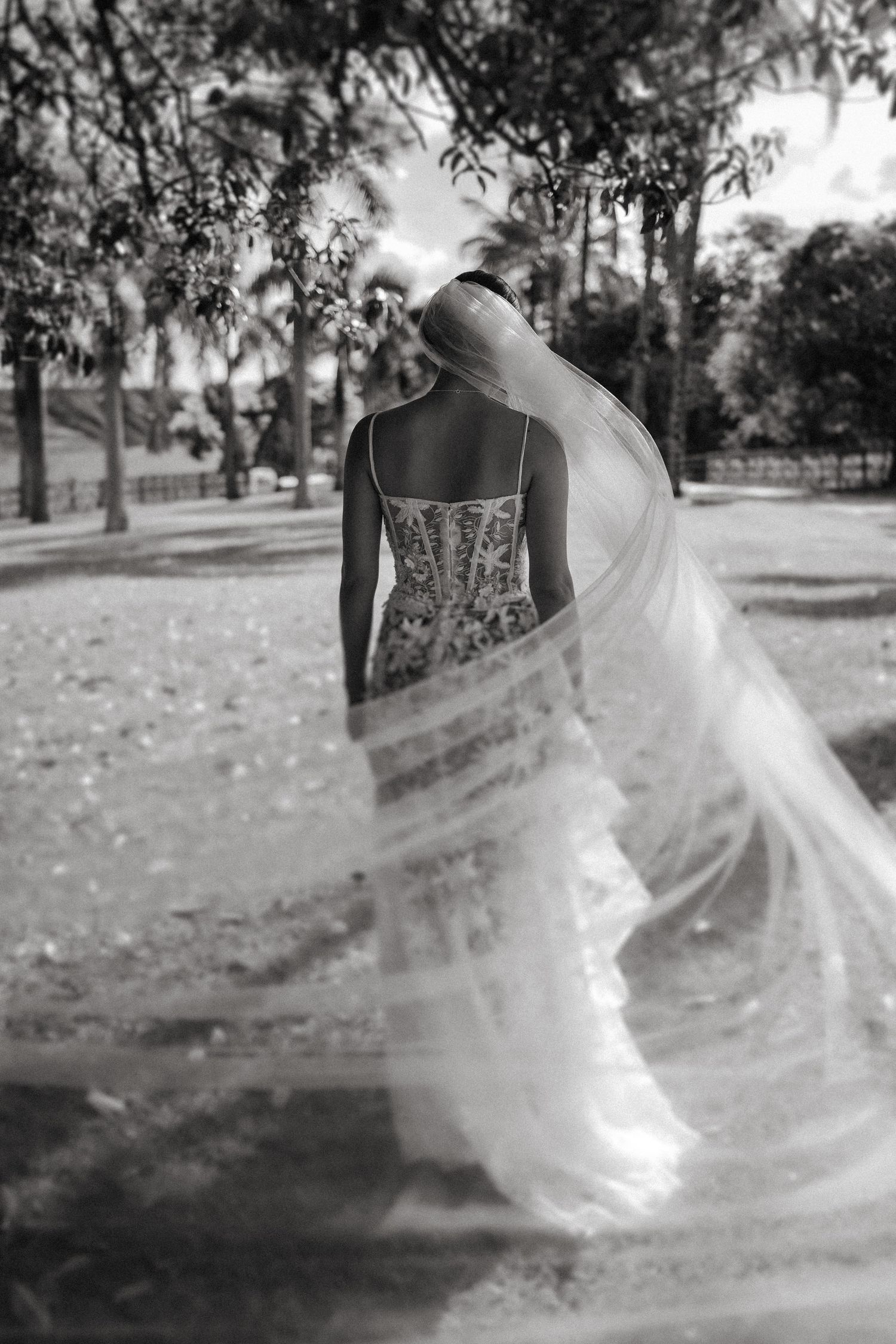 A flowing white wedding dress veil catches the wind in a dreamy outdoor garden setting, creating an ethereal effect.