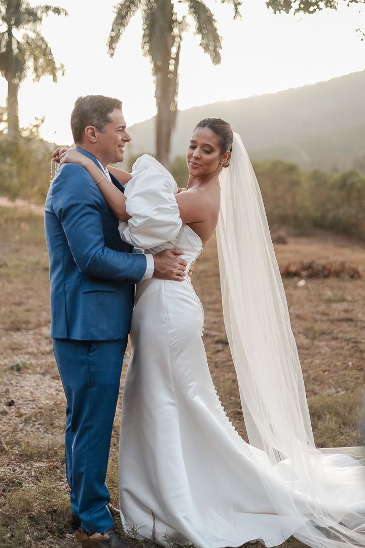 A newlywed couple embraces outdoors at sunset with palm trees and mountains in the background.