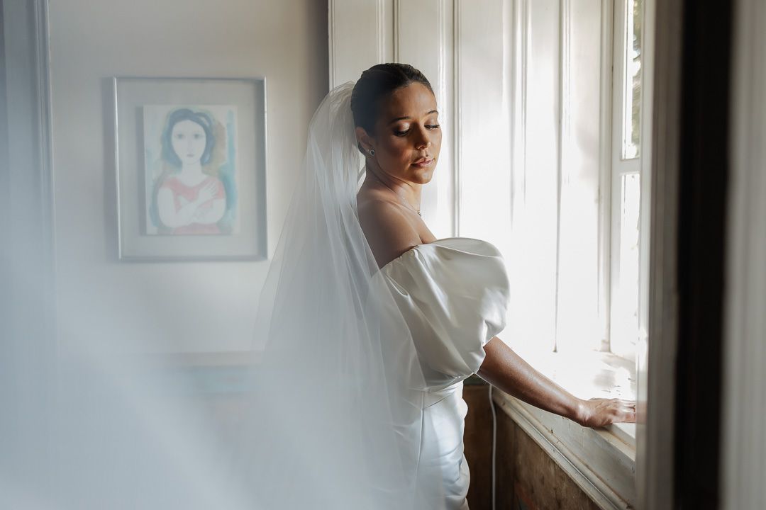 A bride in an elegant white wedding dress poses dreamily by a window with soft natural light filtering in.