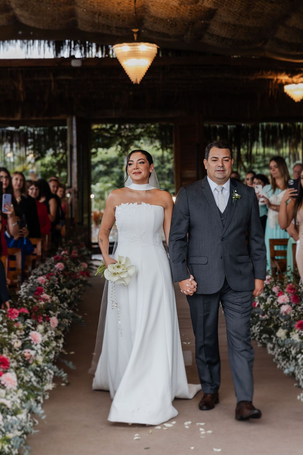 Inspire-se com o casamA couple walks down a flower-lined aisle at their wedding ceremony under a rustic covered venue with pendant lighting.