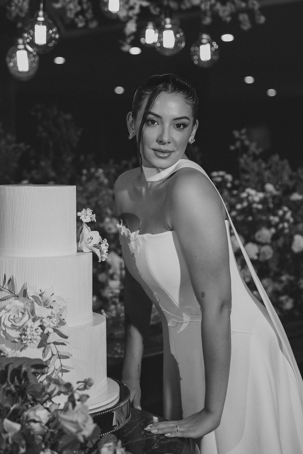 A black and white photograph of someone in a strapless wedding gown standing beside an elegant white tiered cake.
