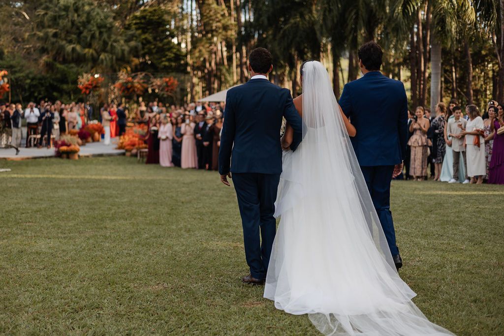 A bride in a flowing white dress walks between two men in navy suits at an outdoor wedding ceremony.