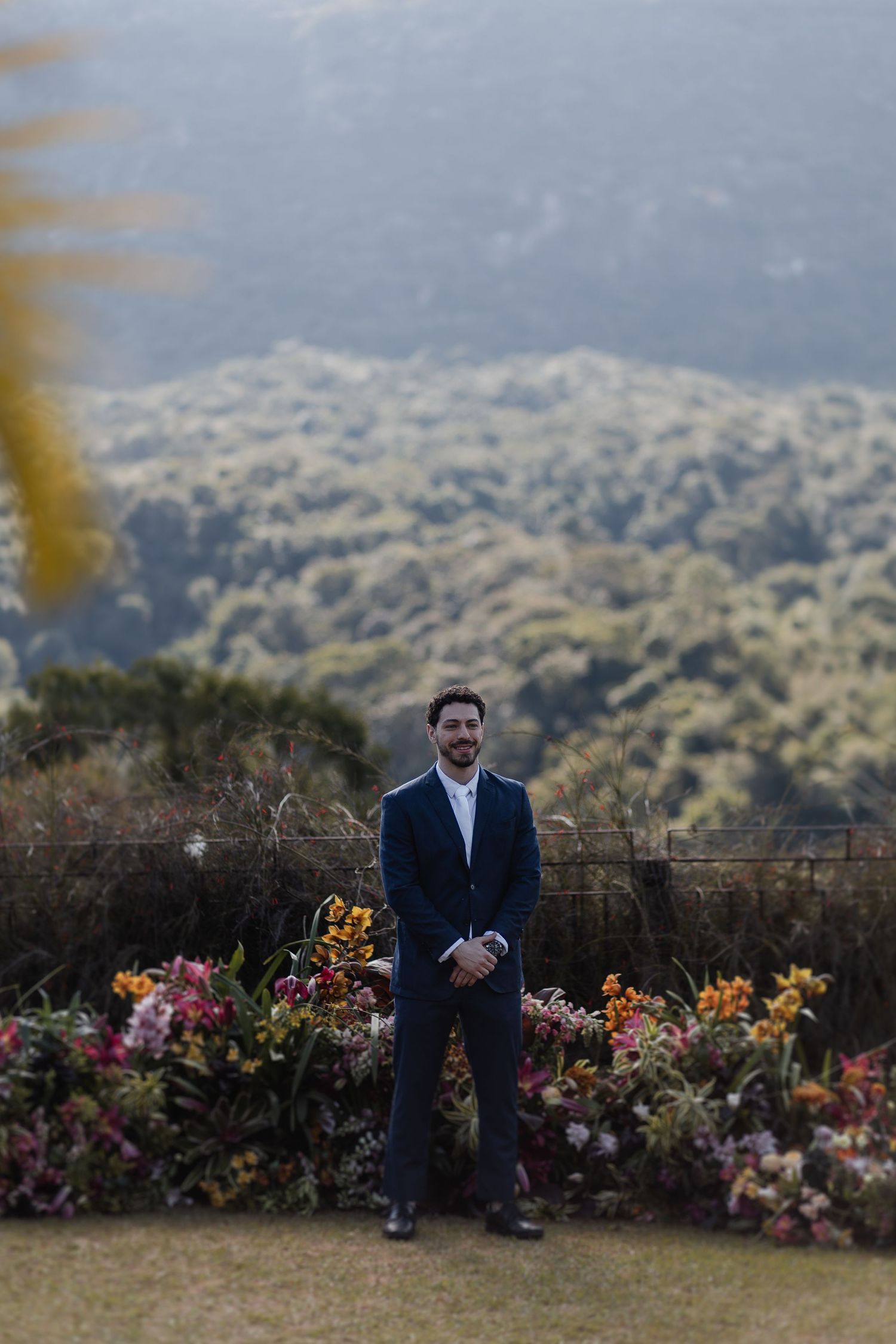 Person in navy suit sits on stone wall surrounded by colorful flowers with mountain landscape in background.