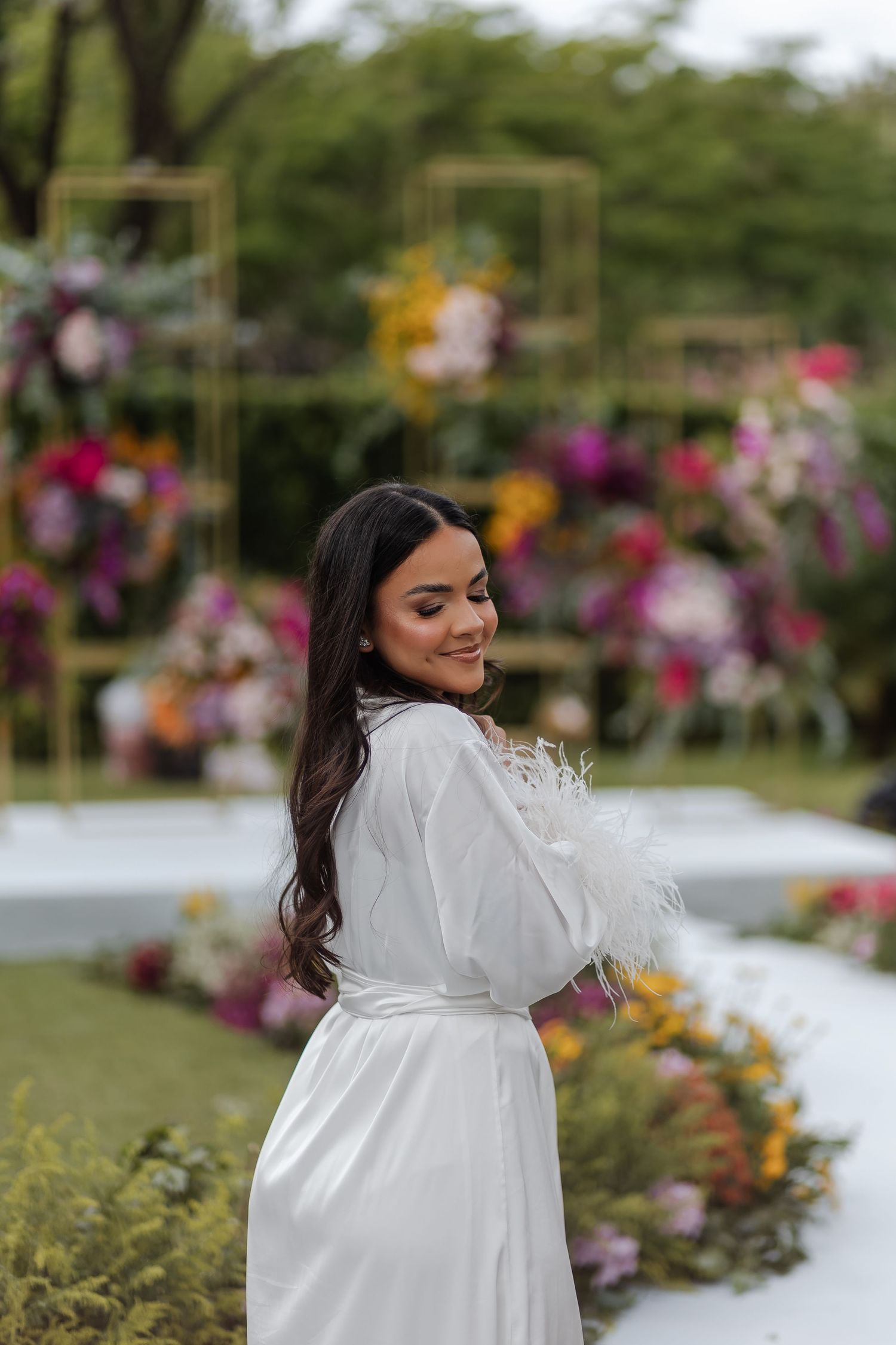 A woman in a white robe dress stands in a garden setting with colorful floral decorations in the background.