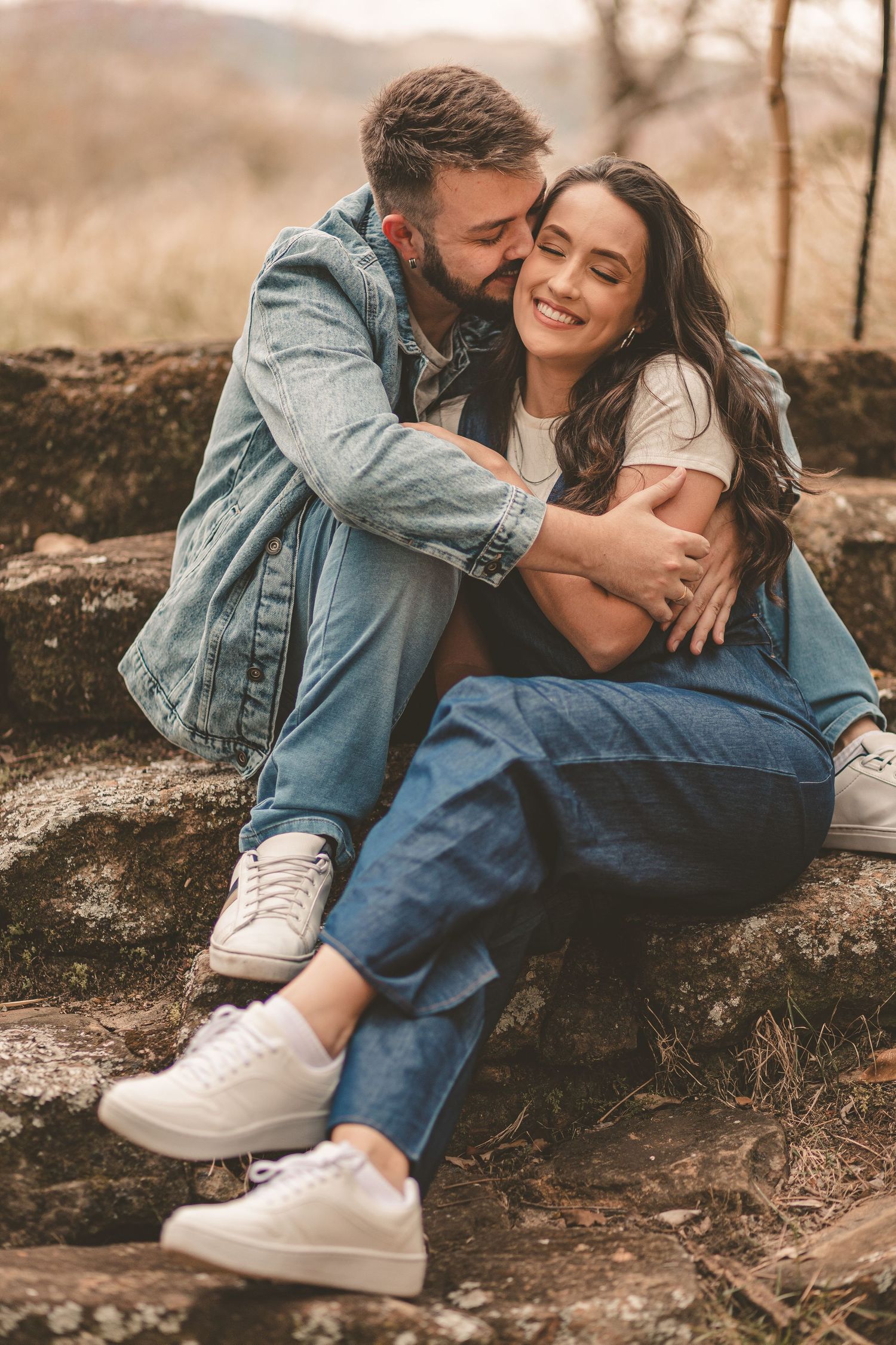 Couple in casual denim outfits embracing on rocks in warm autumn tones during outdoor photoshoot.