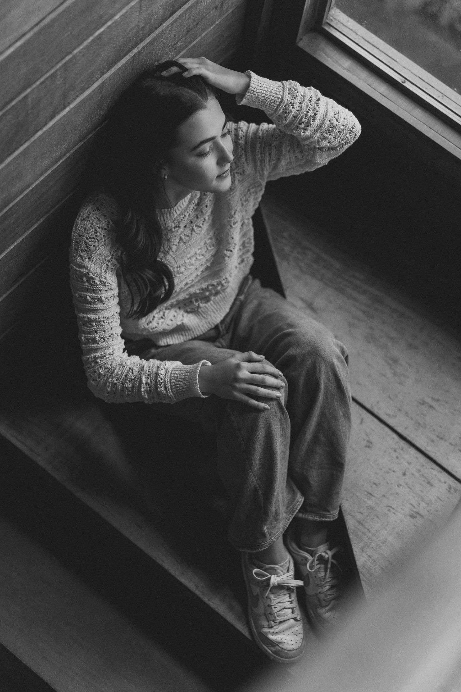 Black and white portrait of person in lace sweater sitting thoughtfully by window in casual pose.