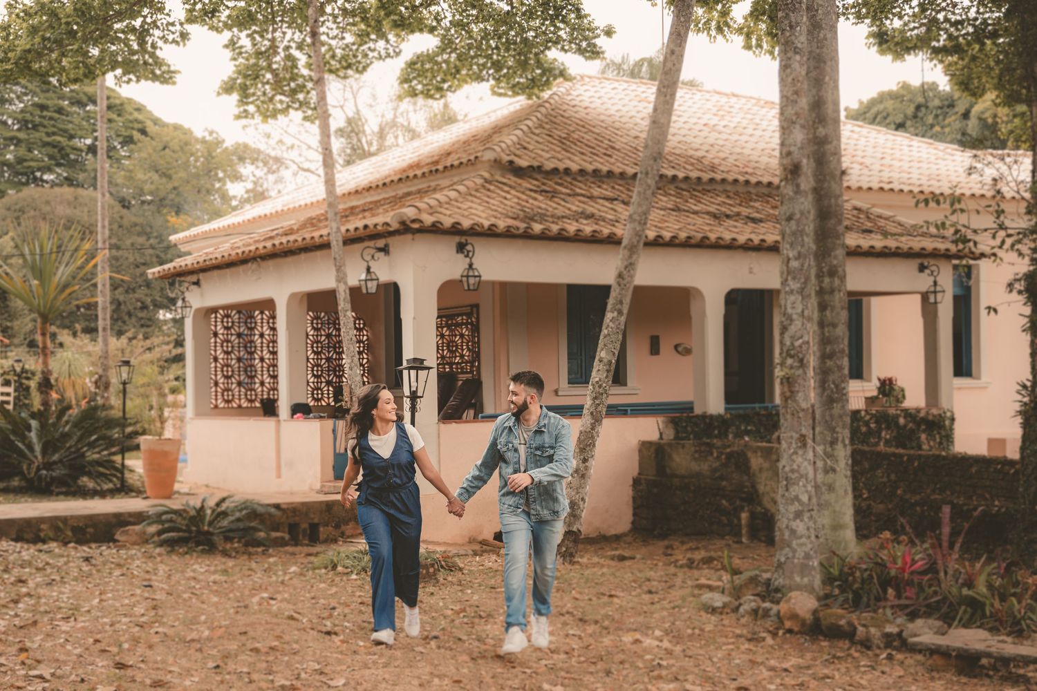 A couple walks hand in hand in front of a pink Mediterranean-style house with terra cotta roof tiles in a wooded setting.