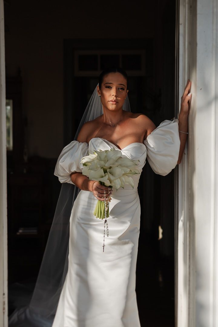 A bride in an elegant white off-shoulder wedding dress holds a bouquet of white flowers while standing in a doorway.