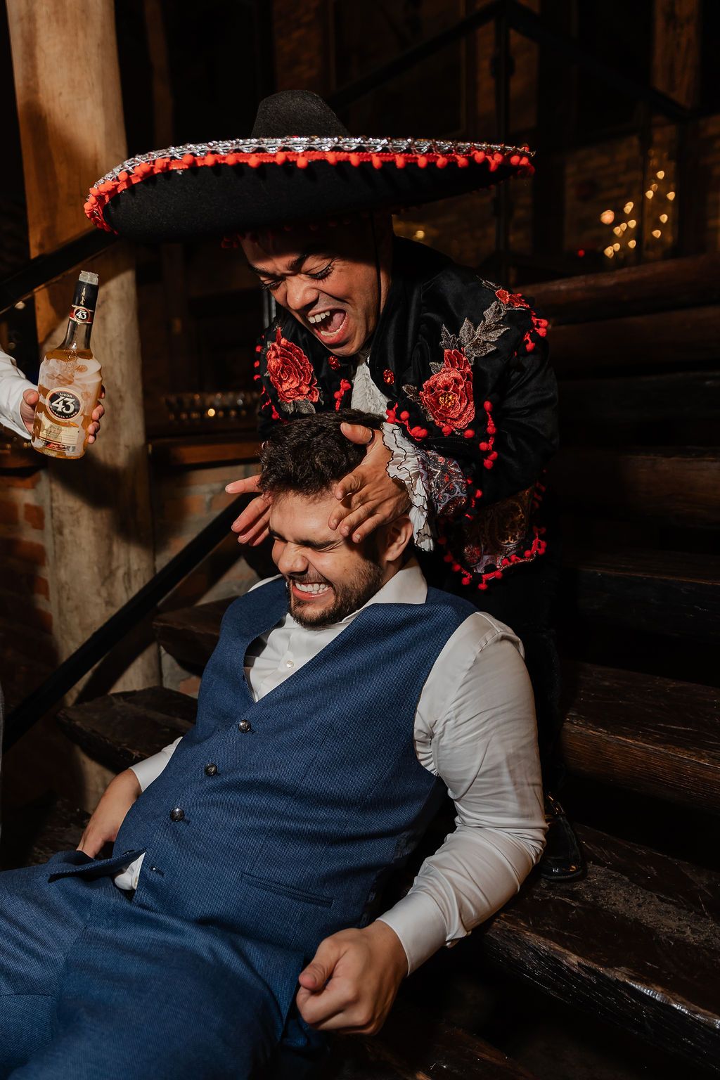 A mariachi performer in traditional hat and costume shares a joyful moment with a guest on wooden stairs.
