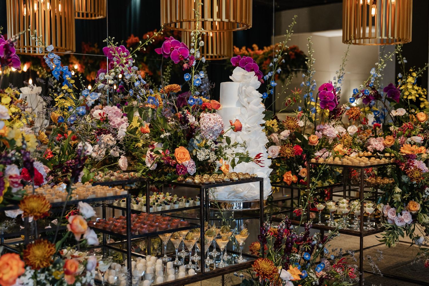 A luxurious wedding dessert table display with a white tiered cake surrounded by colorful floral arrangements and treats.