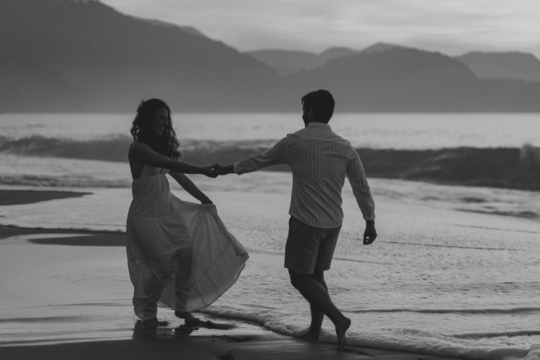 A silhouetted couple holding hands while walking together on a misty beach at sunset with mountains in the background.