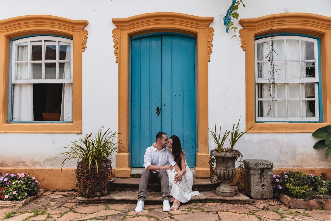 Couple sits on stone steps in front of white building with turquoise door and orange trim in colonial architecture style.
