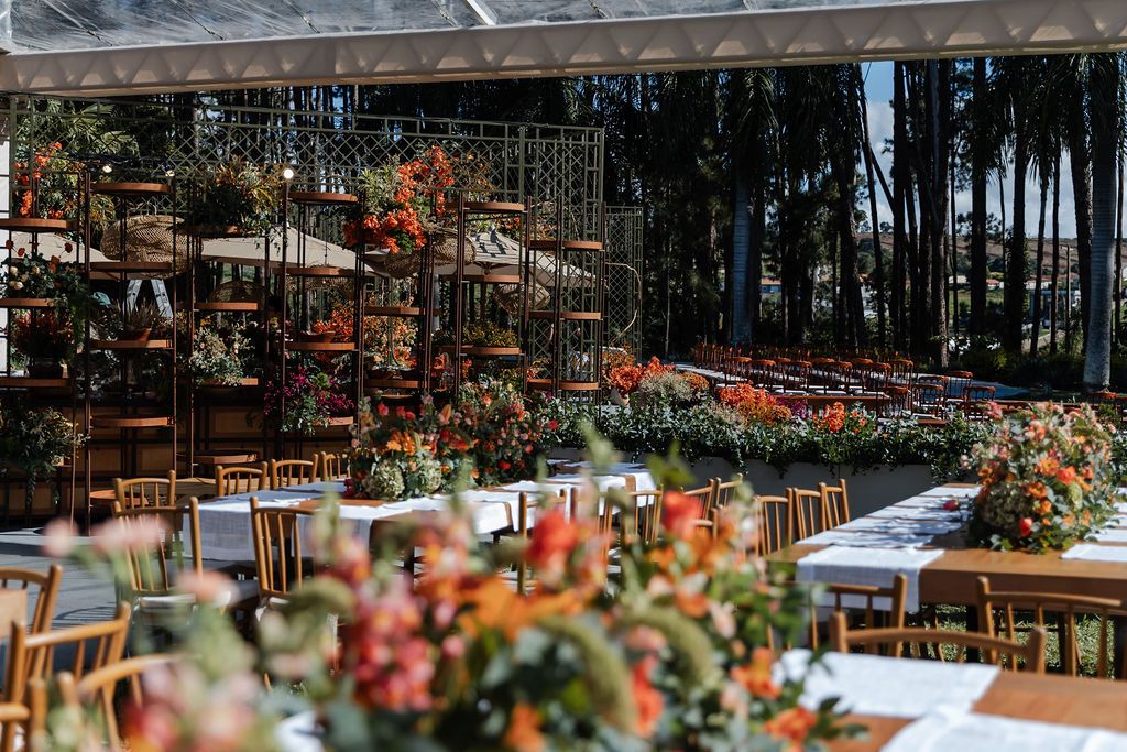 Outdoor wedding reception setup with wooden tables and vibrant coral flower arrangements under a glass-covered venue.