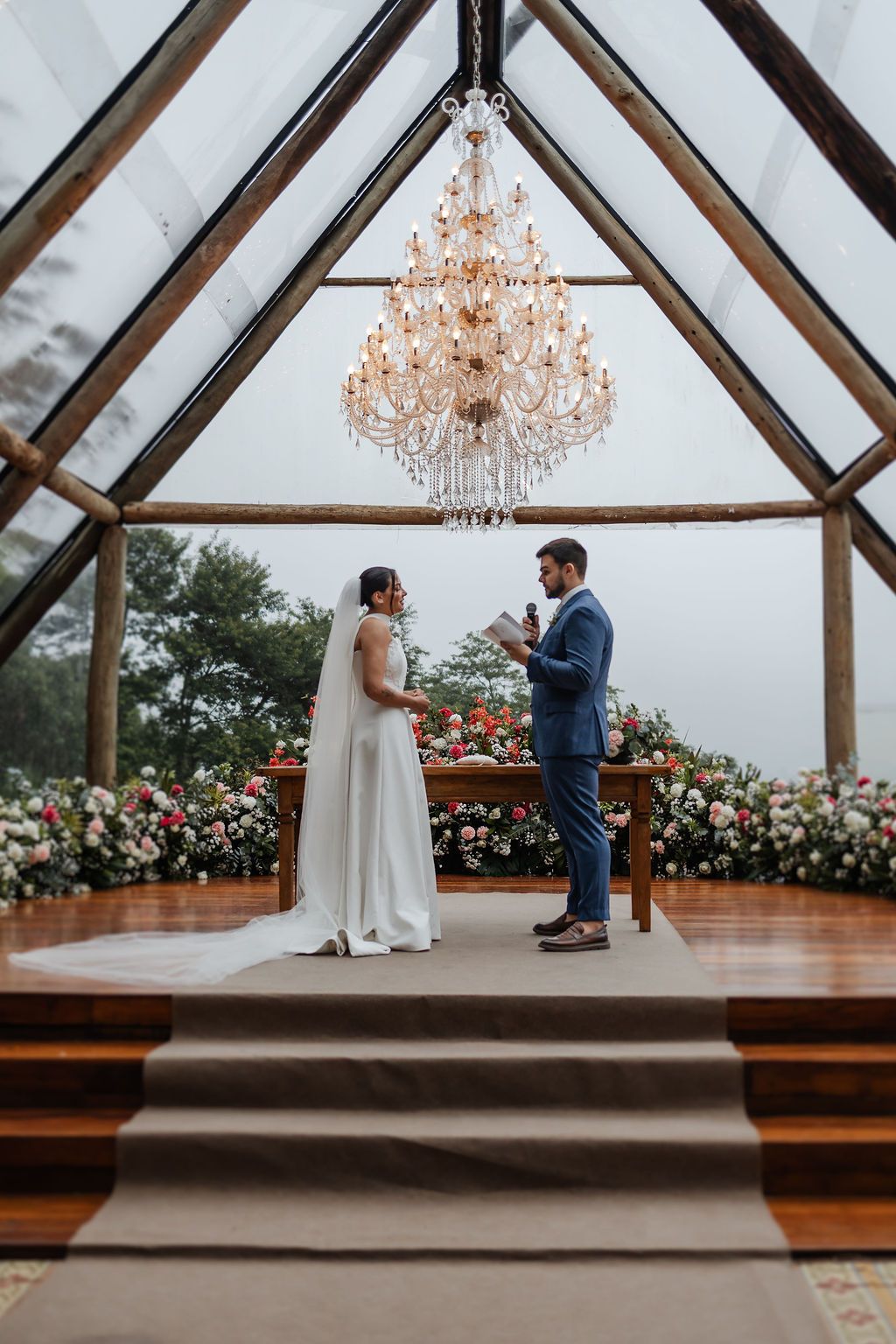 Wedding ceremony in a glass chapel with wooden beams, crystal chandelier, and floral decorations during an outdoor ceremony.