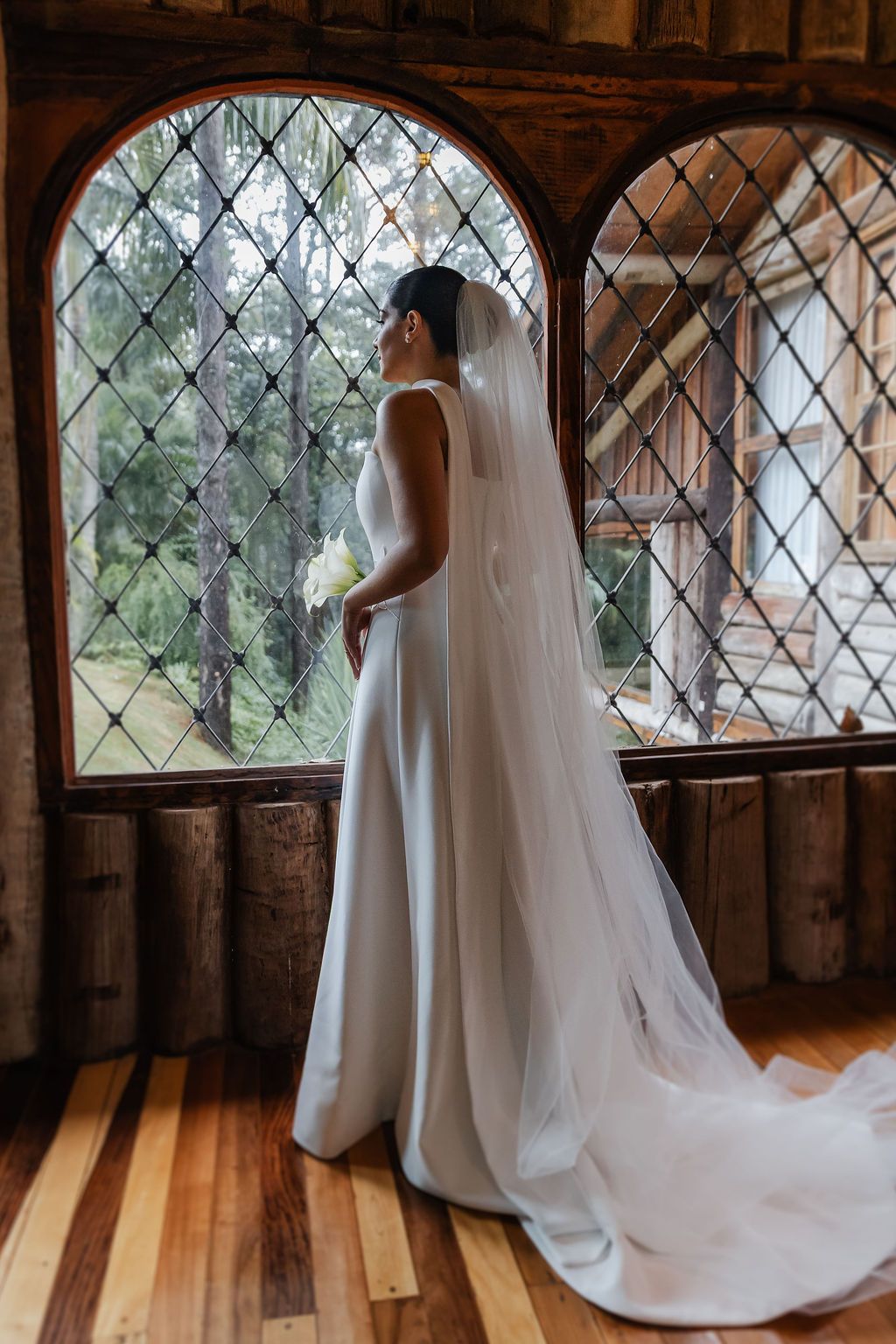 Fotógrafo de casamento em SP, Alfenas e VarginElegant white wedding dress with flowing veil silhouetted in arched window of wooden interior.