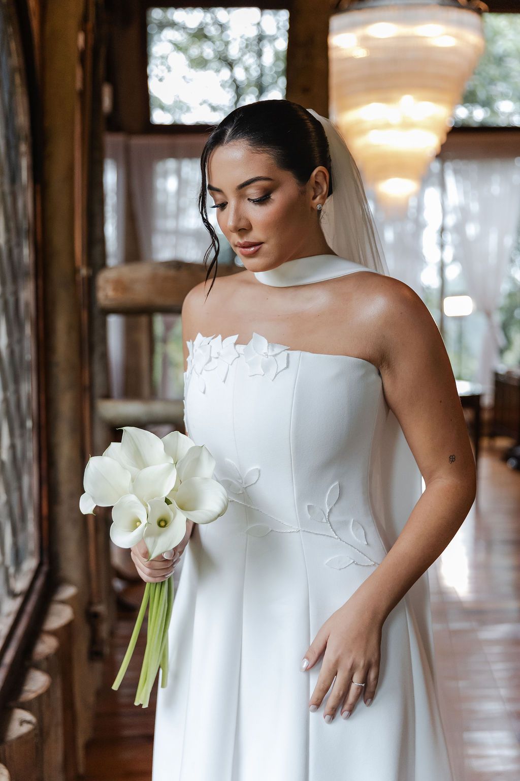 A bride in an elegant white strapless gown with lace details holds a bouquet of white calla lilies in a rustic indoor setting.