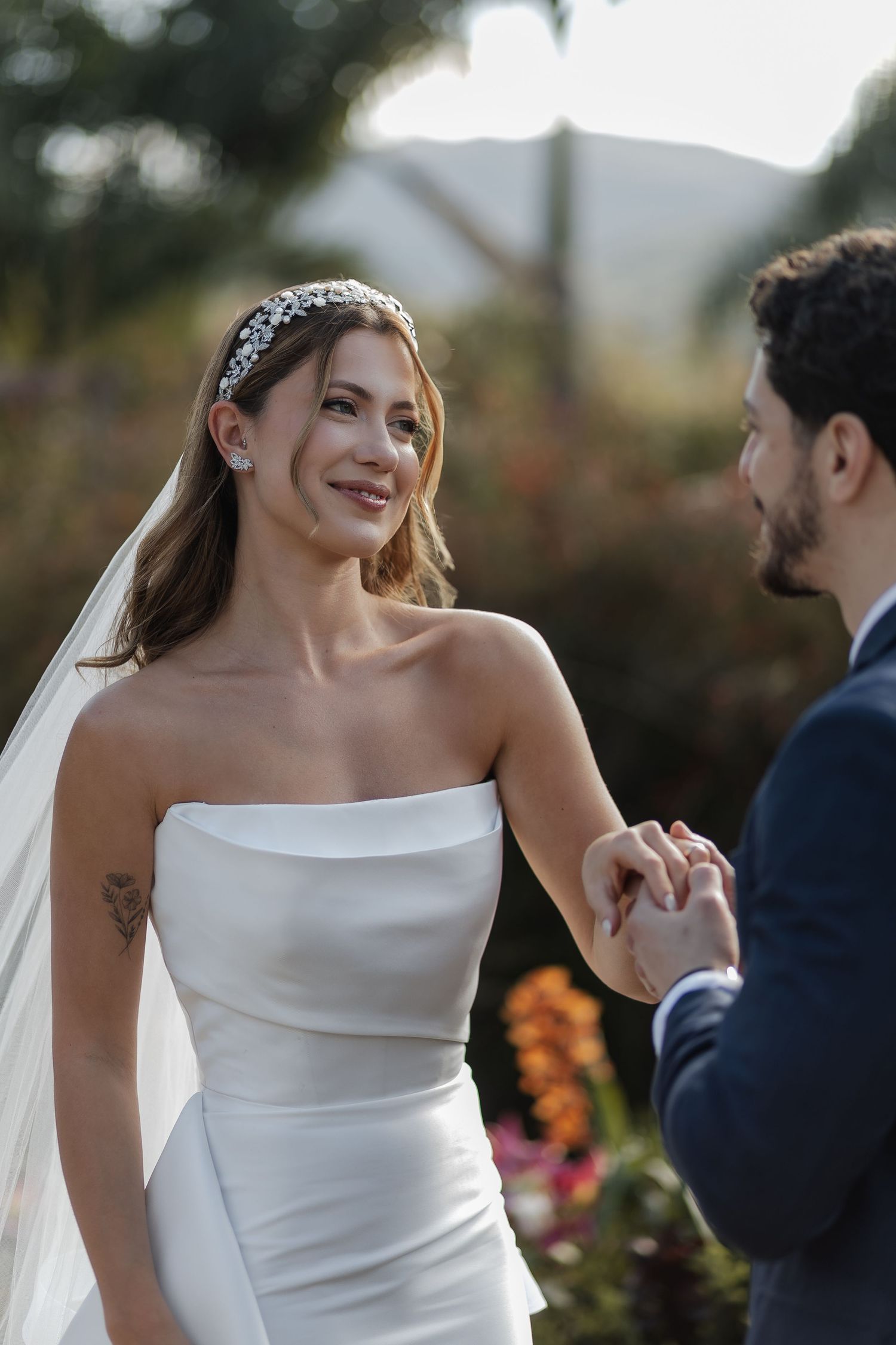 A bride in an elegant off-shoulder wedding dress stands in a garden setting during an outdoor ceremony.